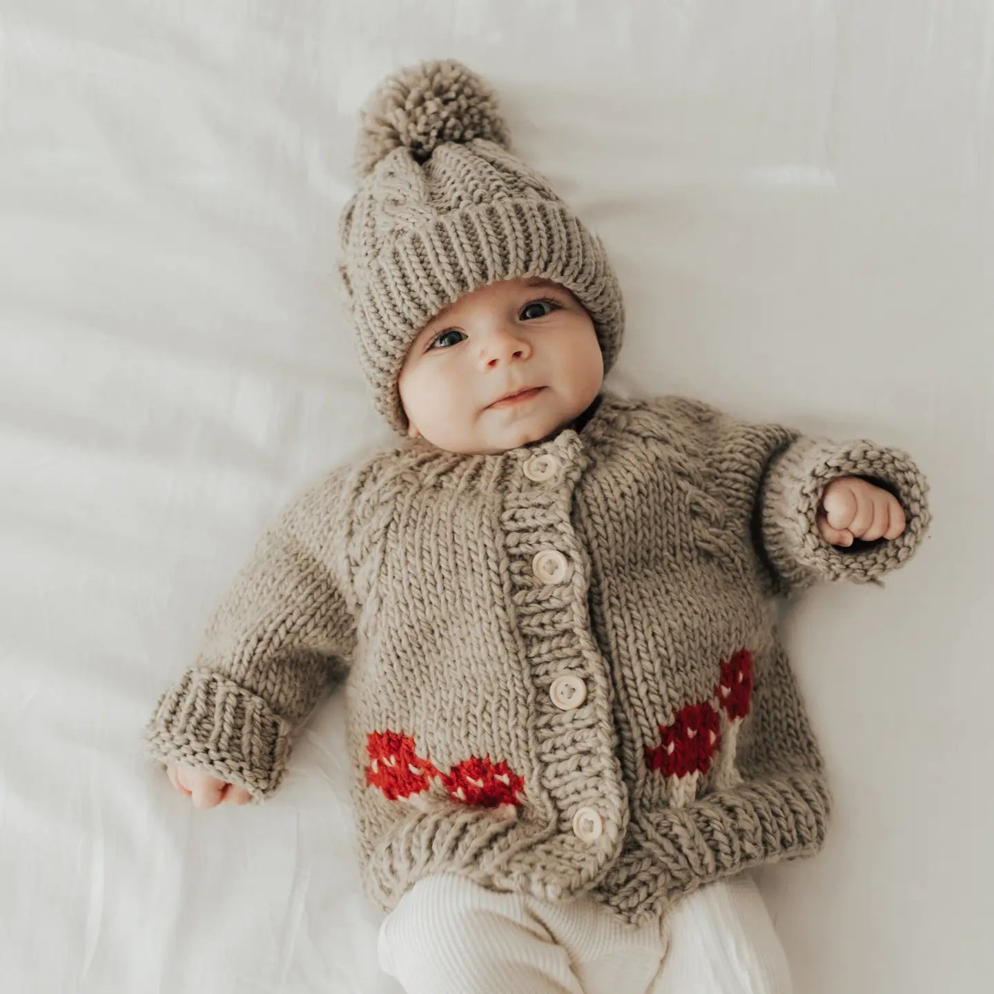 Neutral beige knit baby cardigan with cute red mushroom motif and soft textured yarn.