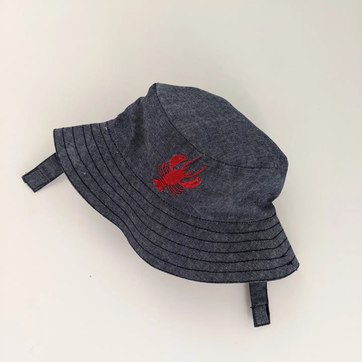 Denim baby bucket hat with red lobster embroidery, adjustable chin strap.