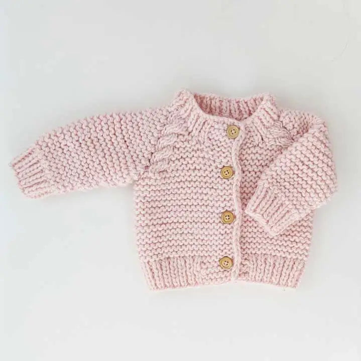 Hand-knitted baby cardigan in soft blush pink yarn with wooden buttons, cozy texture, and ribbed edges.