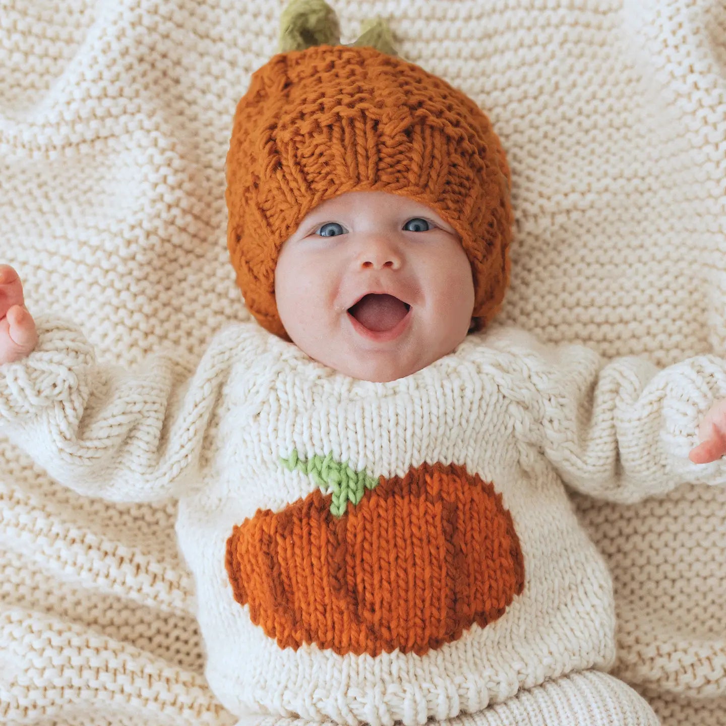 Baby wearing a knitted pumpkin hat and sweater on a soft beige blanket