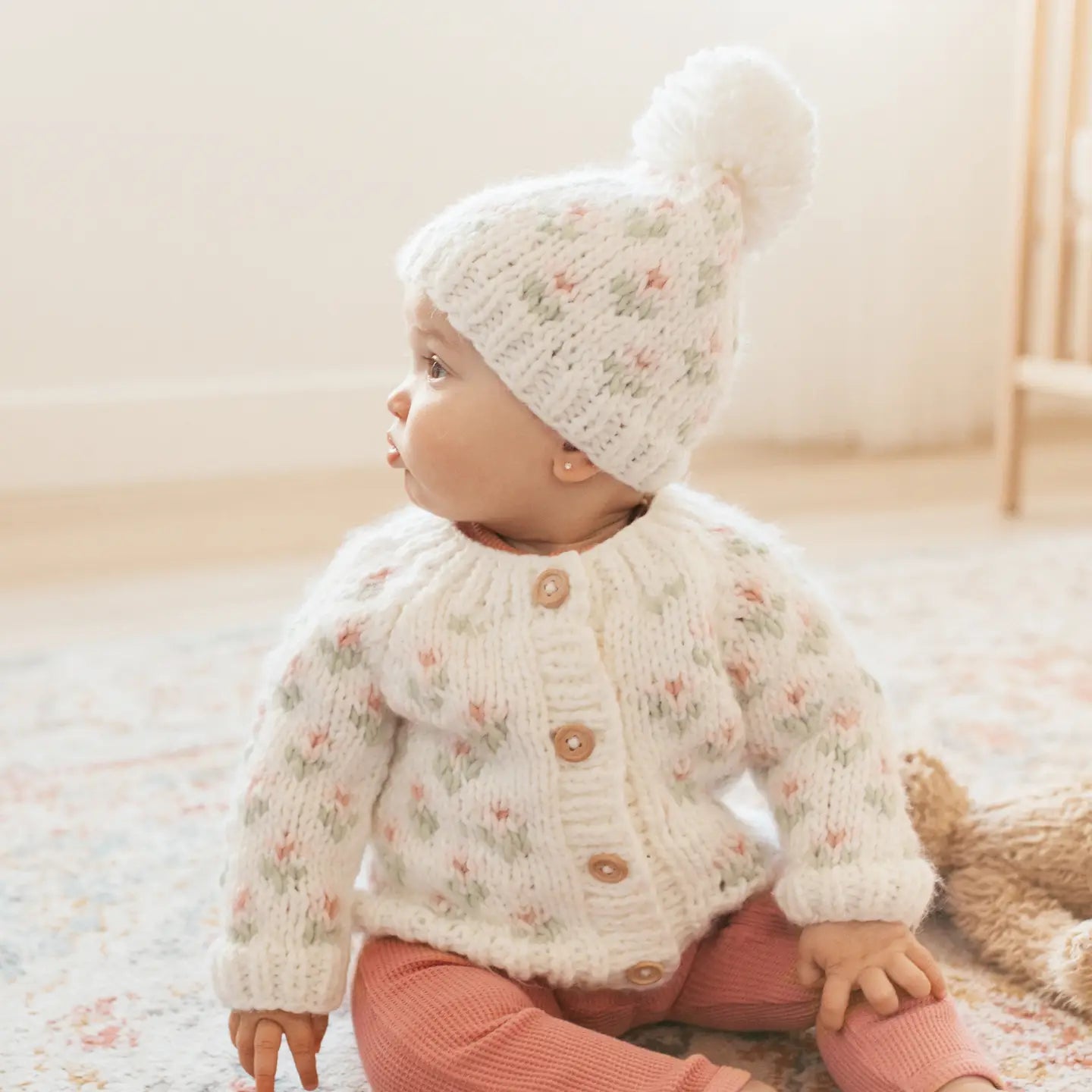 Baby wearing a white knitted cardigan and hat with a fluffy pom-pom, standing on a soft surface.