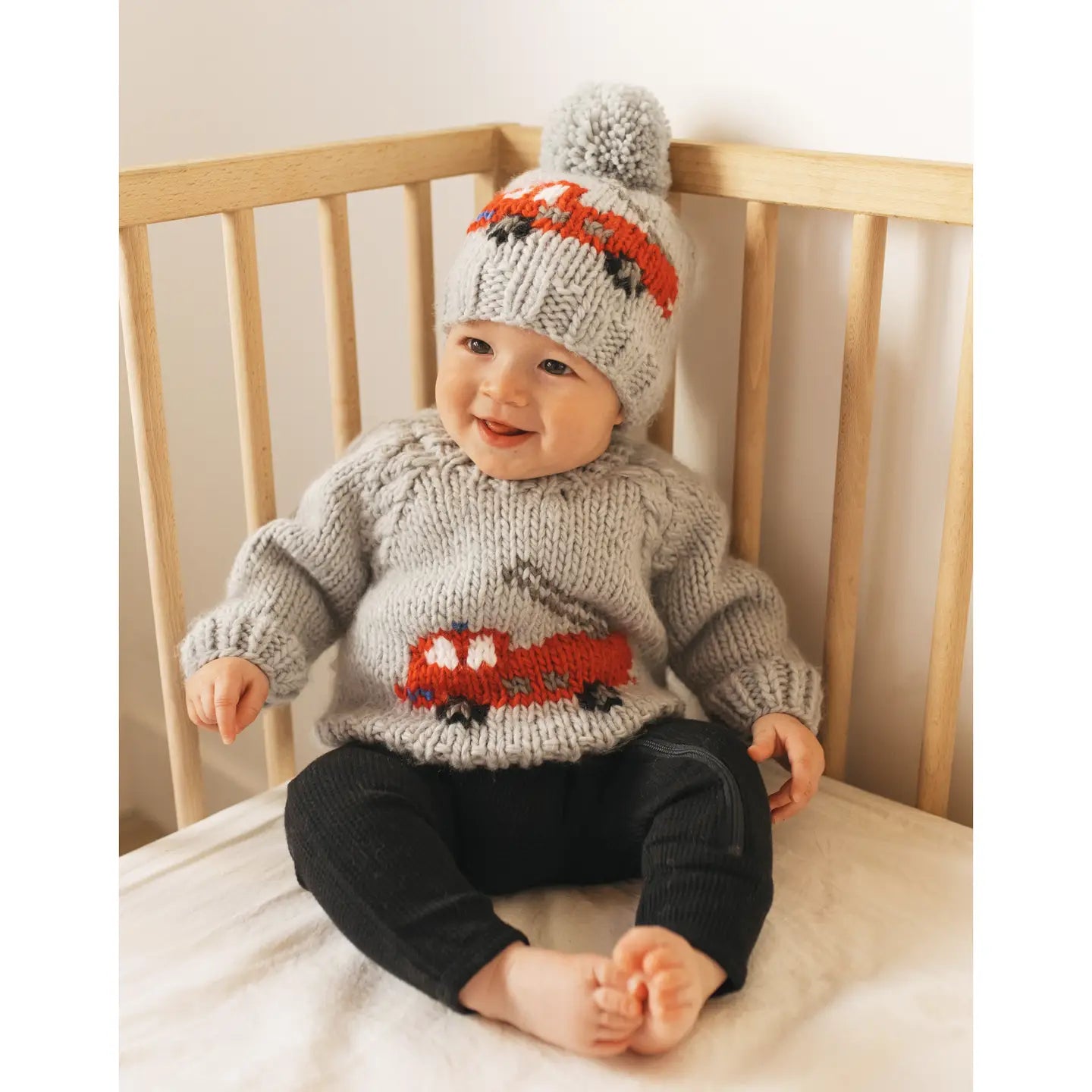 Baby wearing a knitted sweater and hat sitting in a crib.